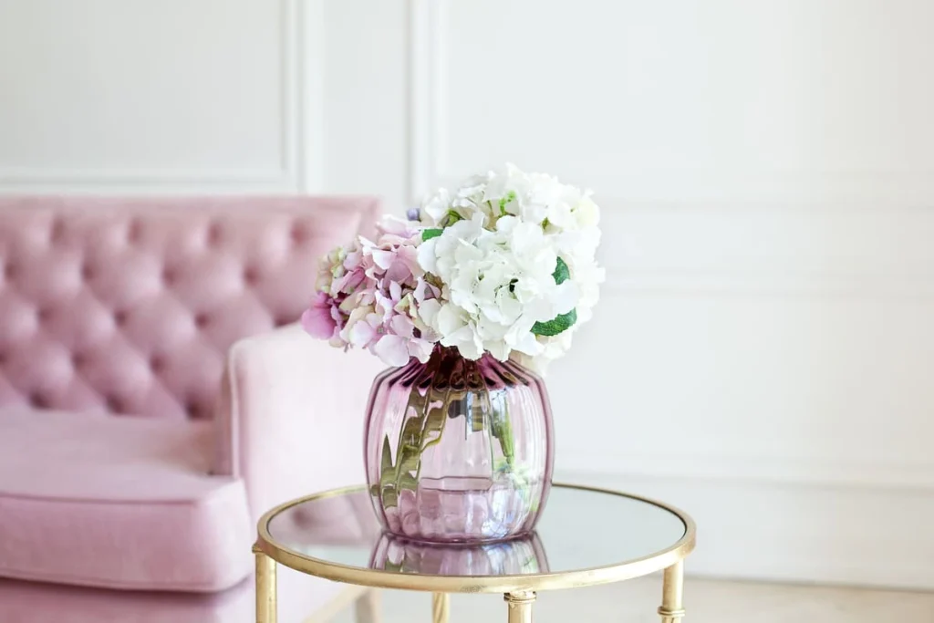 Pastel hydrangeas in a glass vase on a table beside a pink sofa in a white living room