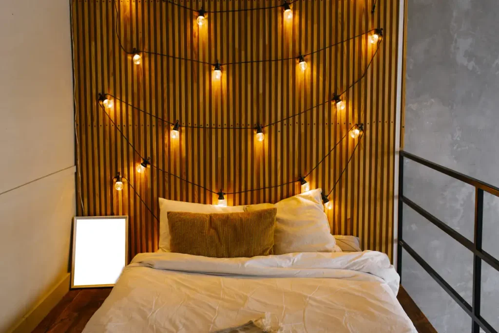 Bed with a wooden slatted headboard lit by warm hanging retro bulbs ·